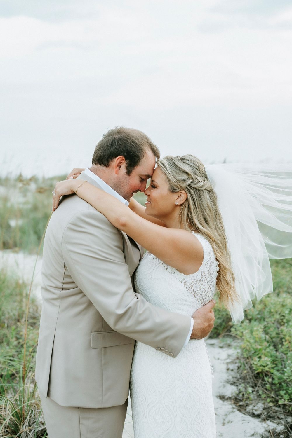 Bride and groom touching foreheads romantically on sandy beach dunes, bride wearing lace gown with flowing veil.