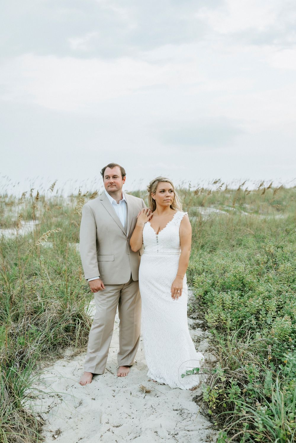 Bride and groom pose barefoot on sandy beach path surrounded by sea oats and green dunes on their wedding day.