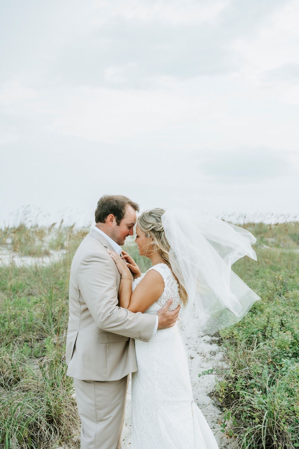 Romantic beach wedding portrait of bride and groom touching foreheads as veil flows in the wind among coastal dunes.
