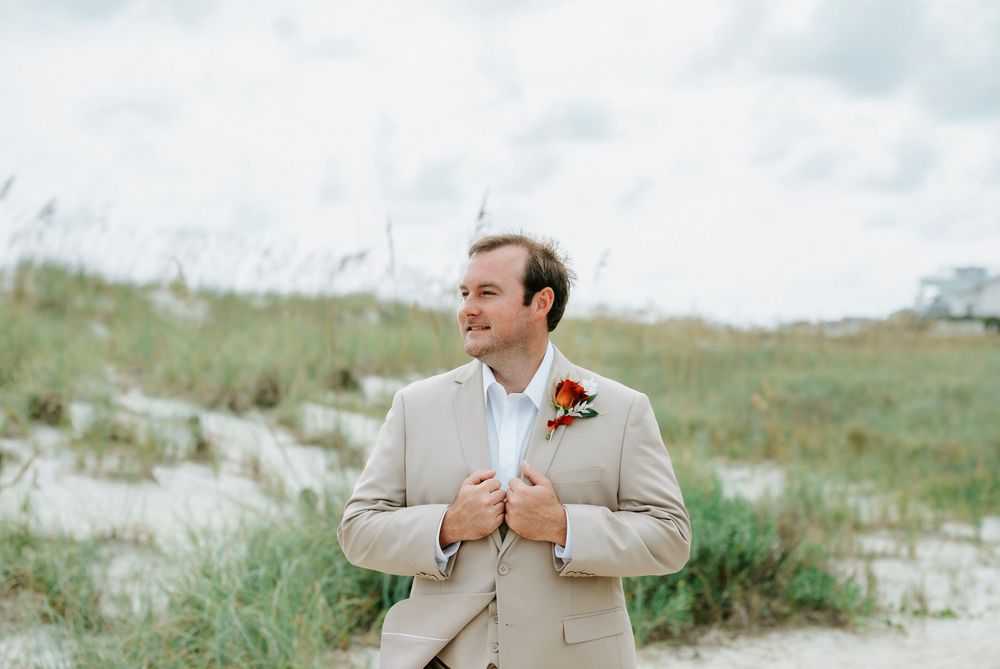 Groom in tan suit with boutonniere adjusting jacket on sandy beach with sea oats in background.