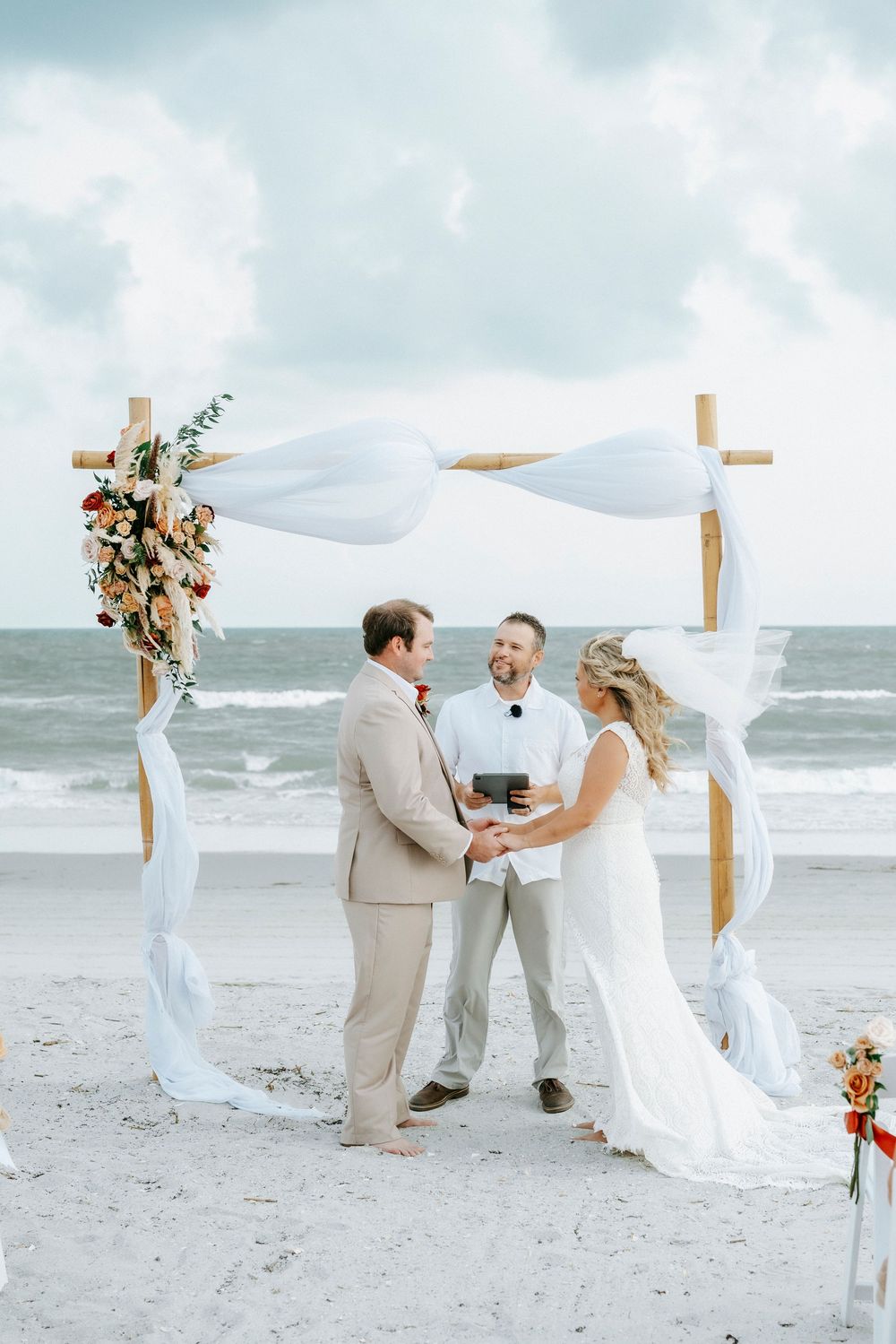 Couple exchanges vows at a beach wedding ceremony under a wooden arch with white draping and floral decor.