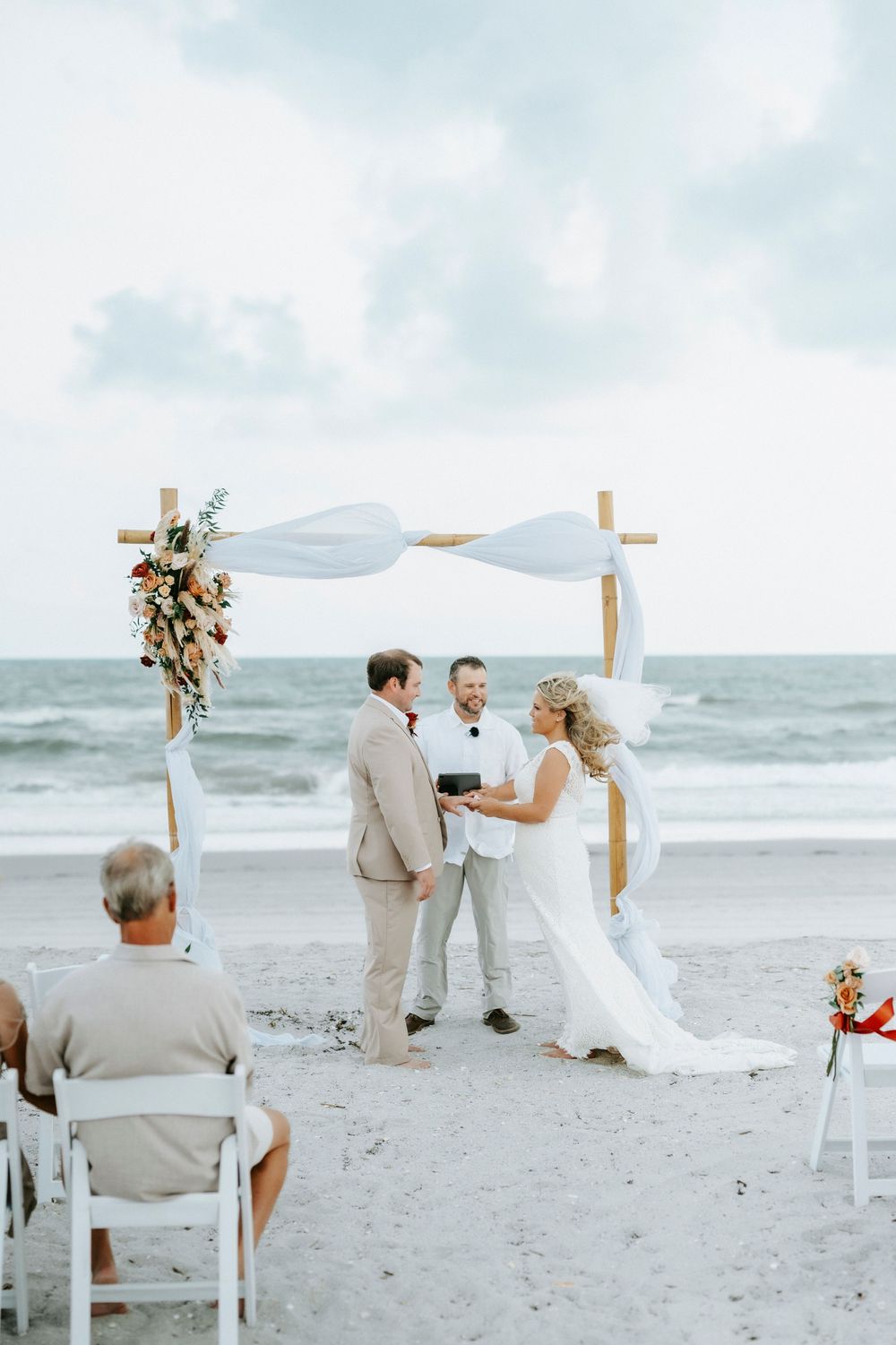 Couple exchanging vows at a romantic beach wedding ceremony under a wooden arch with white draping and floral decor.