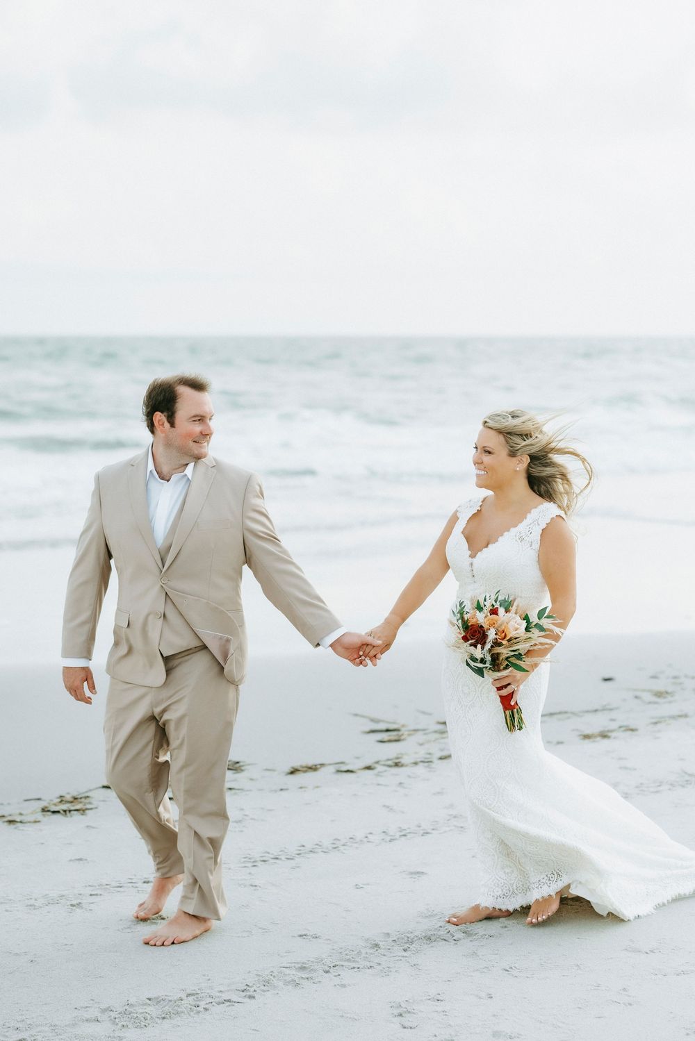 Newlyweds walk hand-in-hand barefoot on a beach, bride in white lace gown holding bouquet, groom in tan suit.