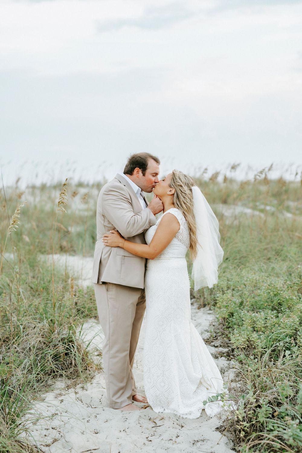 Bride and groom sharing a kiss on sandy beach path surrounded by sea oats, bride in lace gown with veil.
