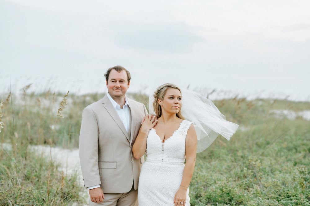 Bride and groom posing outdoors near coastal dunes, bride in white gown with veil, groom in tan suit.