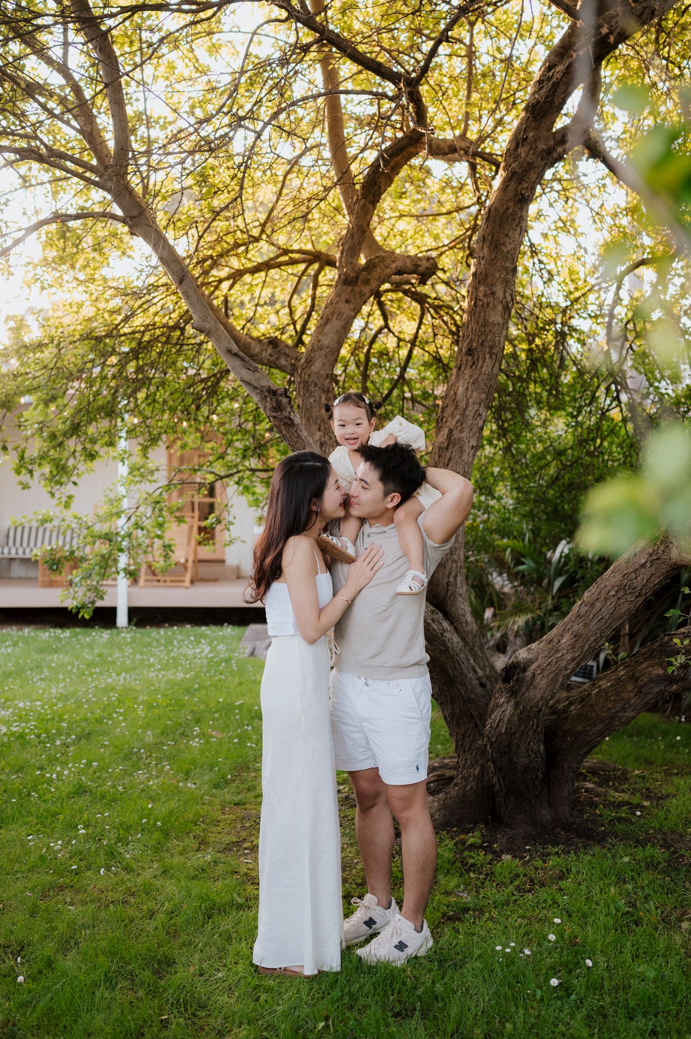 A young family poses under a blooming tree, parents kissing while father holds toddler, dressed in white outfits outdoors.