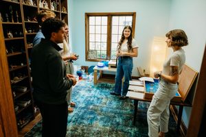 Group of young adults socializing in a cozy room with blue rug, shelves, and natural window light.