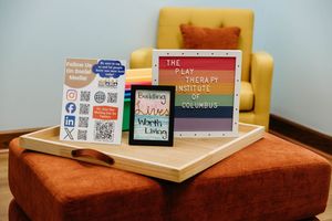 Play Therapy Institute of Columbus display with tablet, book, and QR code sign on wooden tray beside yellow chair.