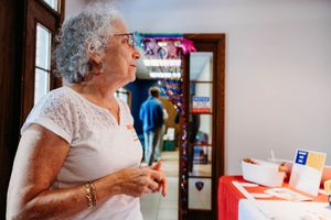 Elderly woman with gray hair in white lace top standing thoughtfully indoors near a decorated table.