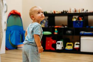 Curious toddler boy standing in a playroom filled with colorful toys on shelves, looking up with wonder.
