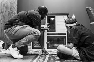 Two people crouching near a small wooden rabbit hutch in a room, examining it in black and white photo.
