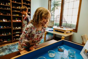 Young girl leaning over a light table with glass bowls in a colorful toy-filled classroom.