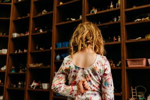 Young girl with curly hair stands facing a large wooden shelf filled with small toys and figurines.