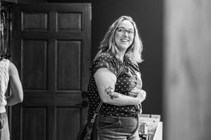 Black and white photo of a smiling woman with glasses, crossed arms, and a tattoo, standing near a wooden door.