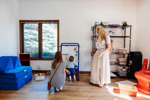 Two women and a child playing in a bright playroom with colorful toys, shelves, and wooden flooring near a window.