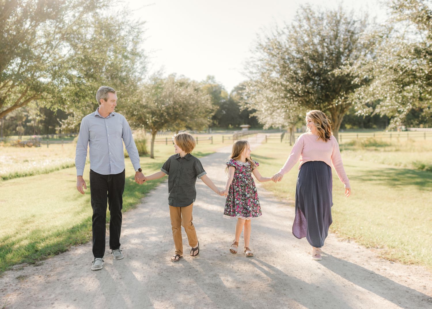 Family of four holding hands walking down a sunny tree-lined path outdoors.