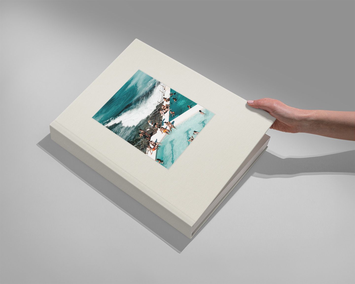 Hand holding a large white photo book with a teal ocean wave and floral collage art on the cover.