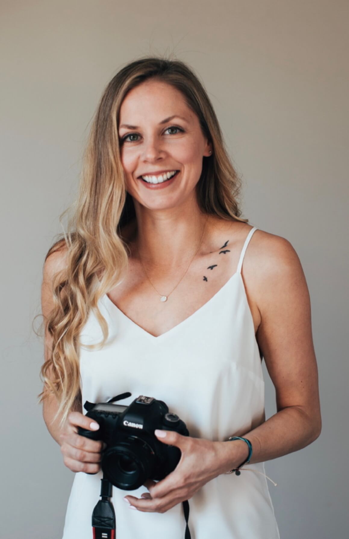 Smiling blonde woman holding a Canon camera wearing a white dress with bird tattoo on chest.