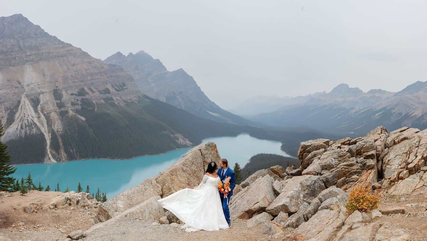 Bride and groom pose on rocky overlook above turquoise Peyto Lake with misty mountain peaks in Banff, Canada.