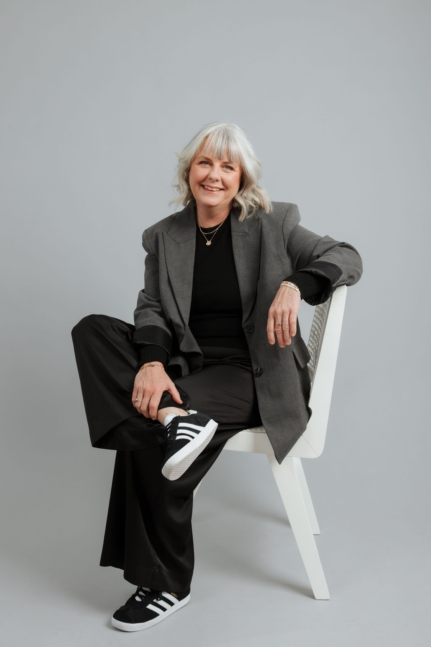 Smiling older woman with gray hair sitting on white chair wearing gray blazer, black pants, and Adidas sneakers.