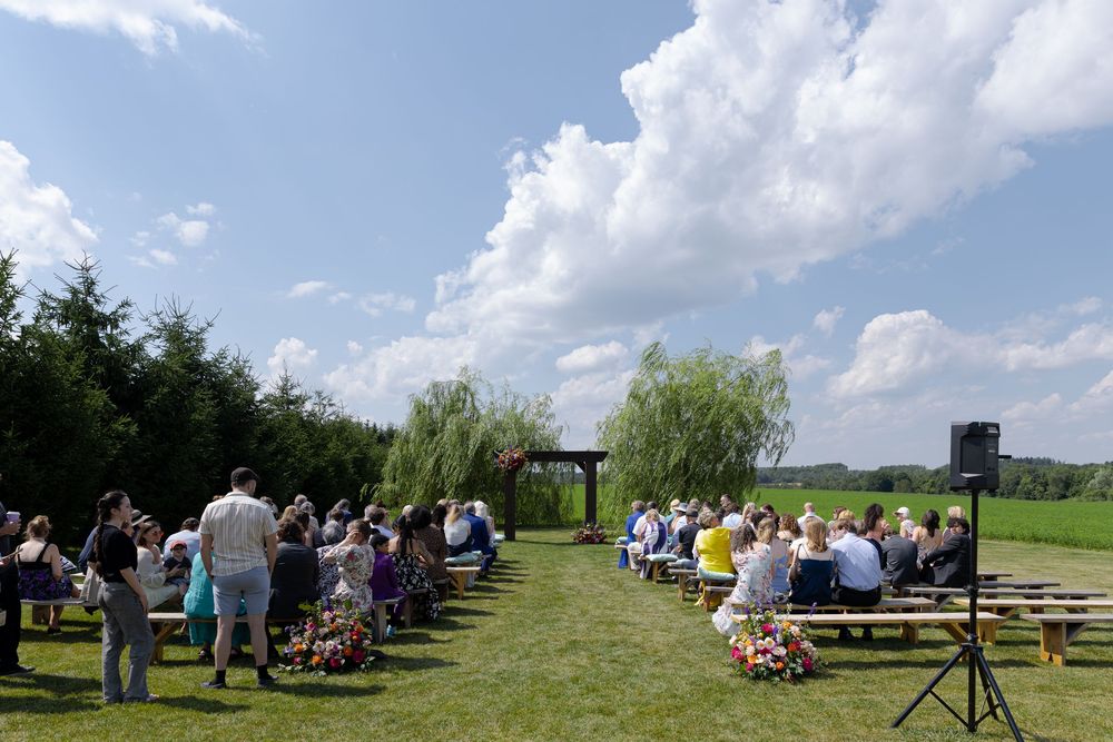 Outdoor wedding ceremony on green lawn with guests seated on wooden benches at barn swallow fields in guelph, ontario.