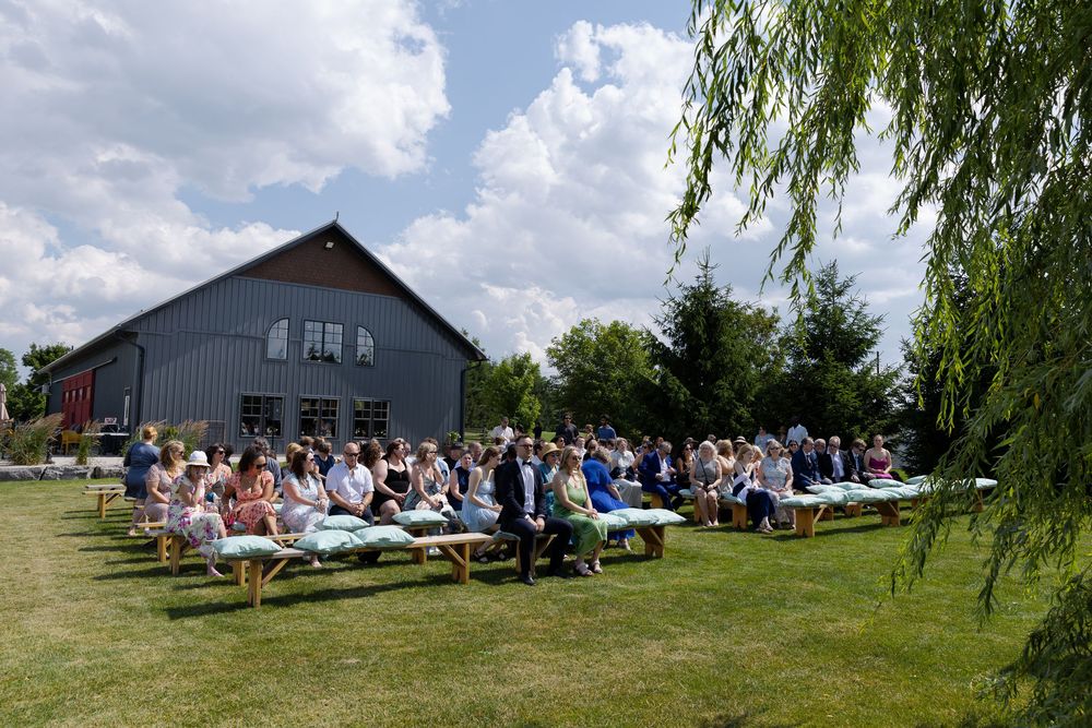 Guests seated on wooden benches at an outdoor wedding ceremony on a sunny day at barn swallow fields in guelph, ontario.
