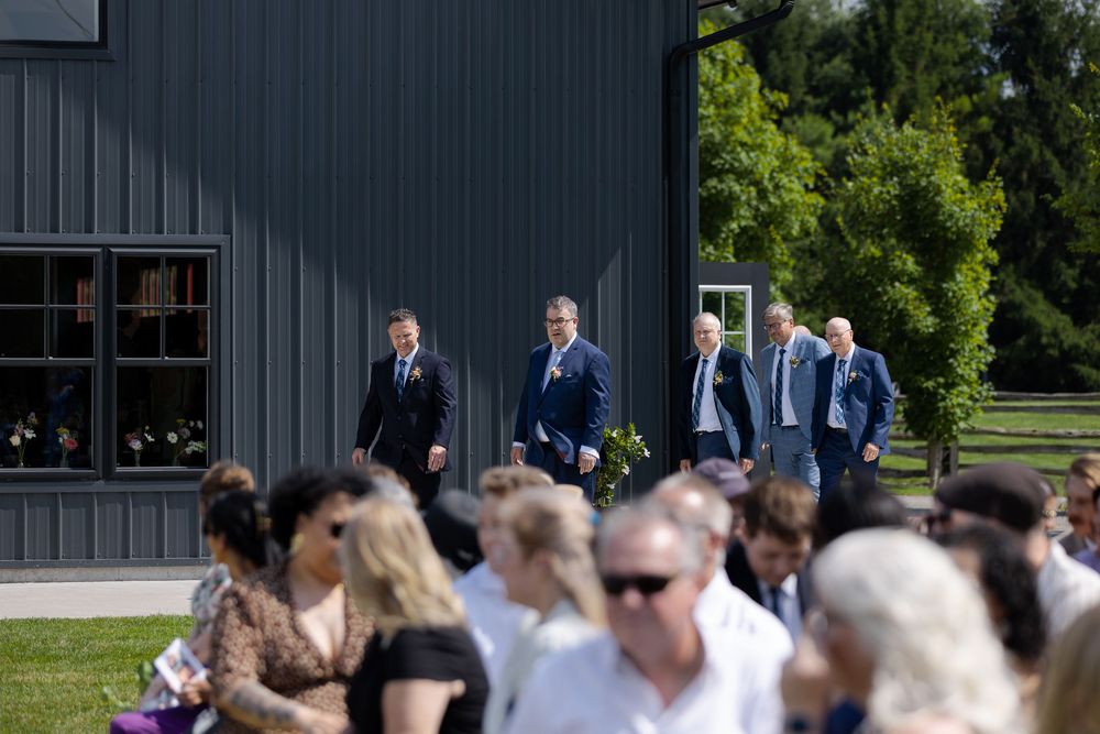 Groomsmen in suits walk toward outdoor wedding guests near a modern dark barn venue on a sunny day at barn swallow fields in guelph, ontario