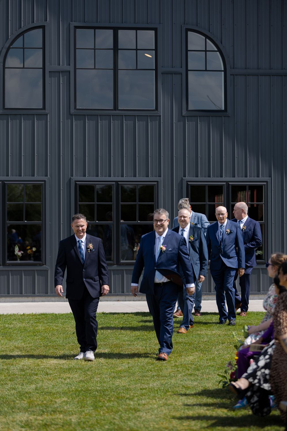 Groomsmen in navy suits walk across green lawn toward outdoor wedding ceremony at barn swallow fields in guelph, ontario.