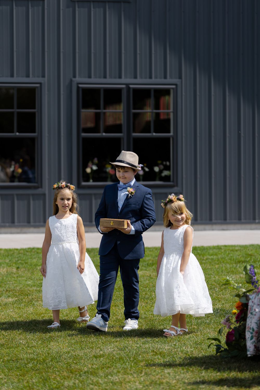 A ring bearer in navy suit and hat walks down the aisle with flower girls in white dresses at barn swallow fields in guelph, ontario.