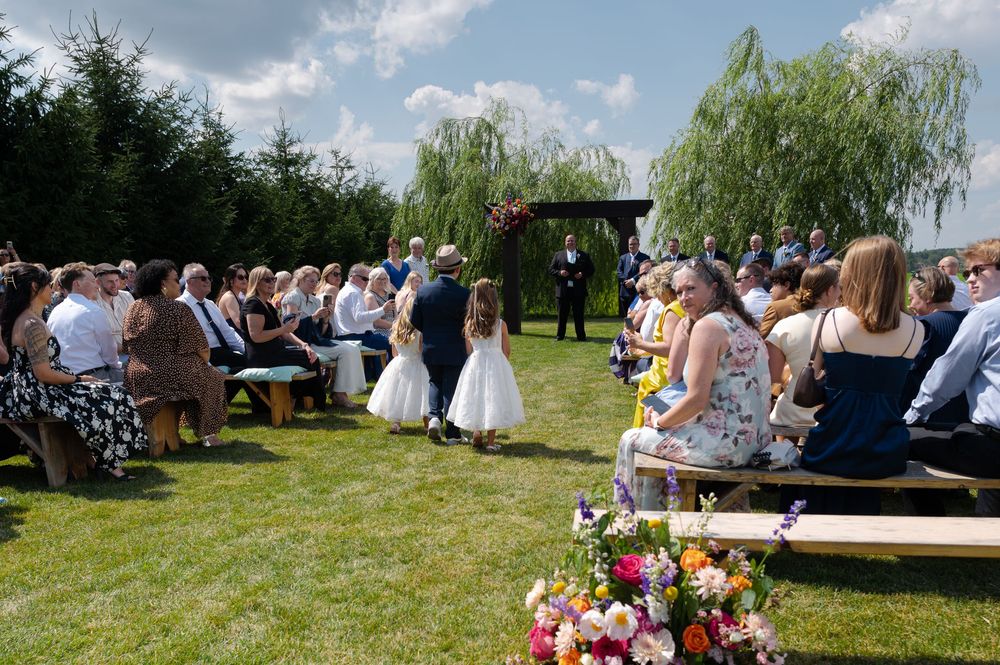 Outdoor wedding ceremony on green lawn with flower girls walking the aisle, guests seated on wooden benches at barn swallow fields in guelph
