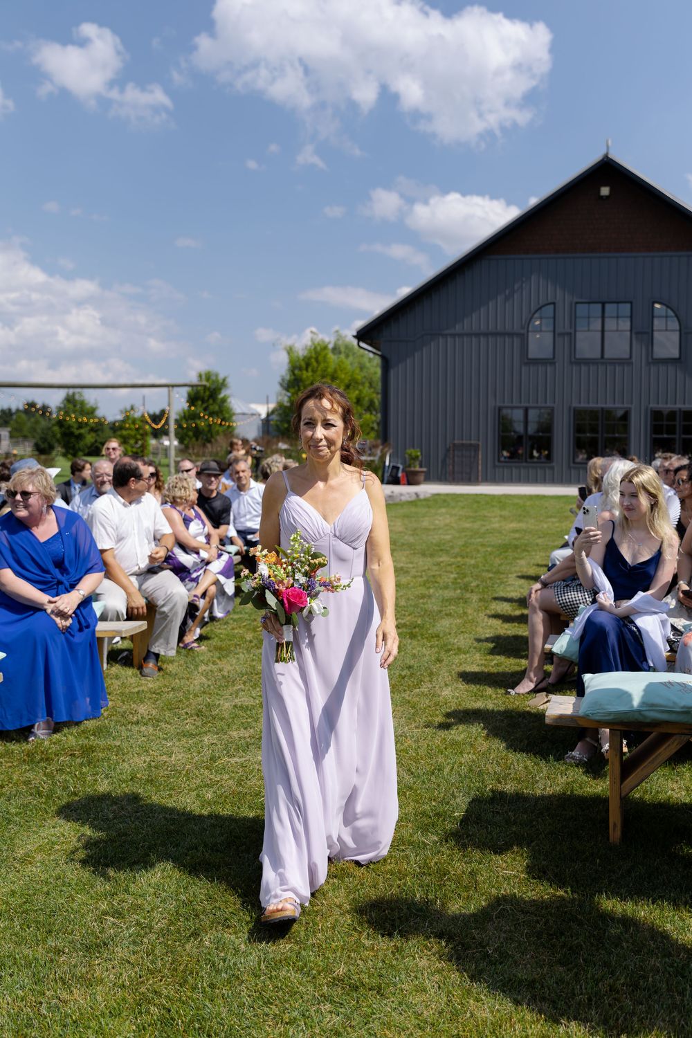 Bridesmaid in lavender dress walks down outdoor wedding aisle at barn swallow fields in guelph, ontario.