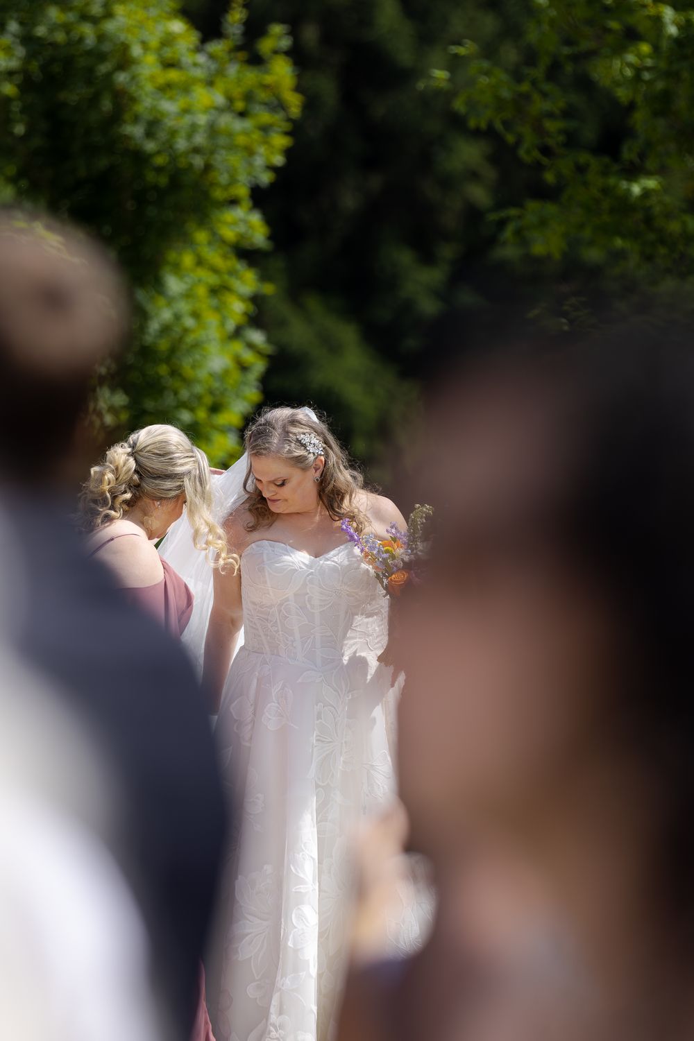 Bride in white lace gown walks down outdoor aisle, bridesmaid adjusting veil, at barn swallow fields in guelph, ontario.