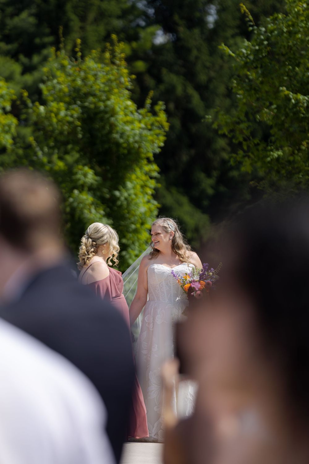Bride stands with bridesmaid in pink dress during outdoor wedding ceremony at barn swallow fields in guelph, ontario.
