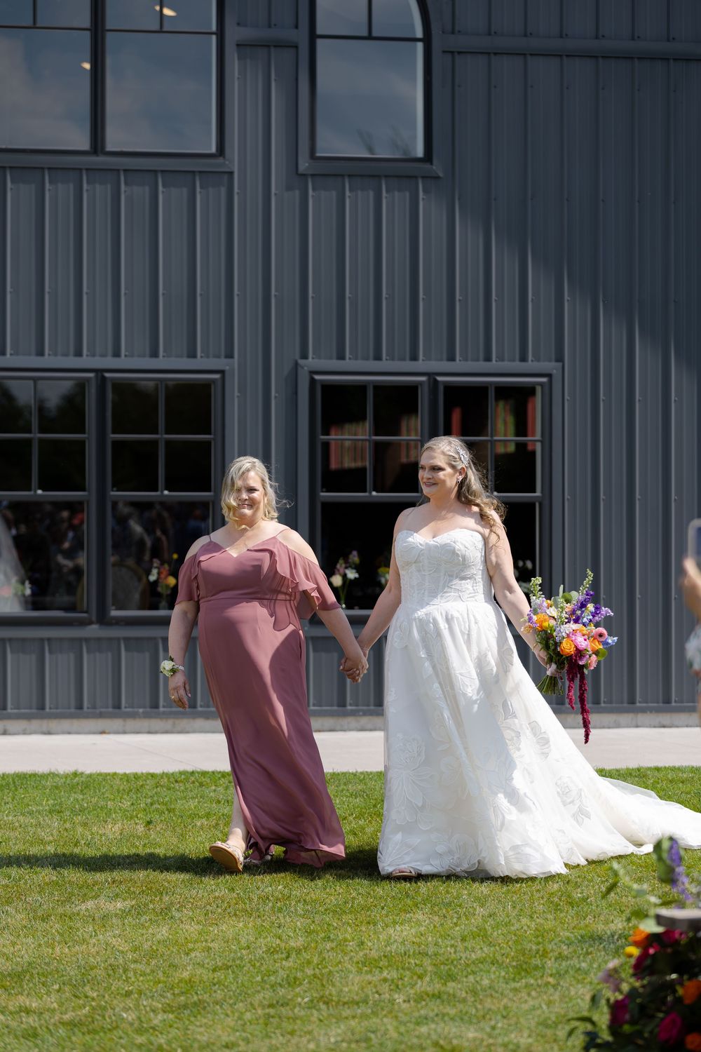Bride in white gown holds colorful bouquet walking with bridesmaid during outdoor ceremony at barn swallow fields in guelph, ontario.