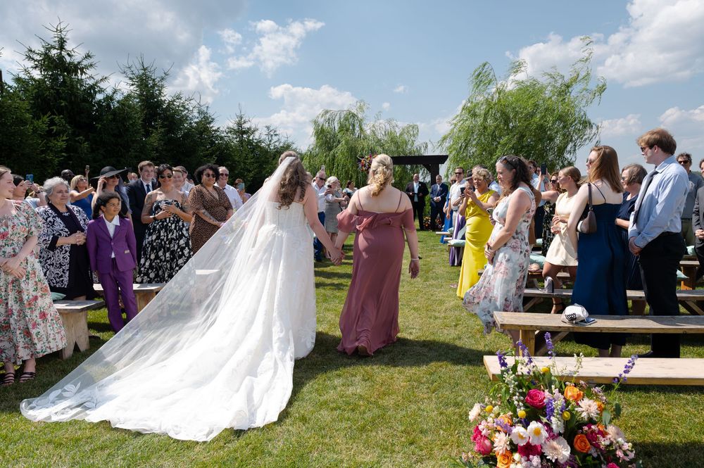 Bride walks outdoor aisle with bridesmaid at sunny garden wedding ceremony at barn swallow fields in guelph, ontario.