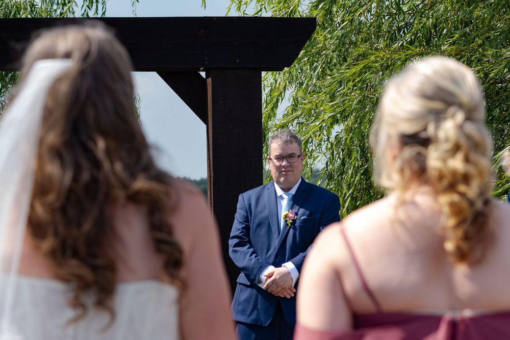 Groom in navy suit watches bride approach at outdoor wedding ceremony at barn swallow fields in guelph, ontario.