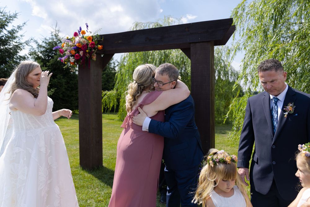 Outdoor wedding ceremony with bridesmaid hugging groom while bride watches at barn swallow fields in guelph, ontario.