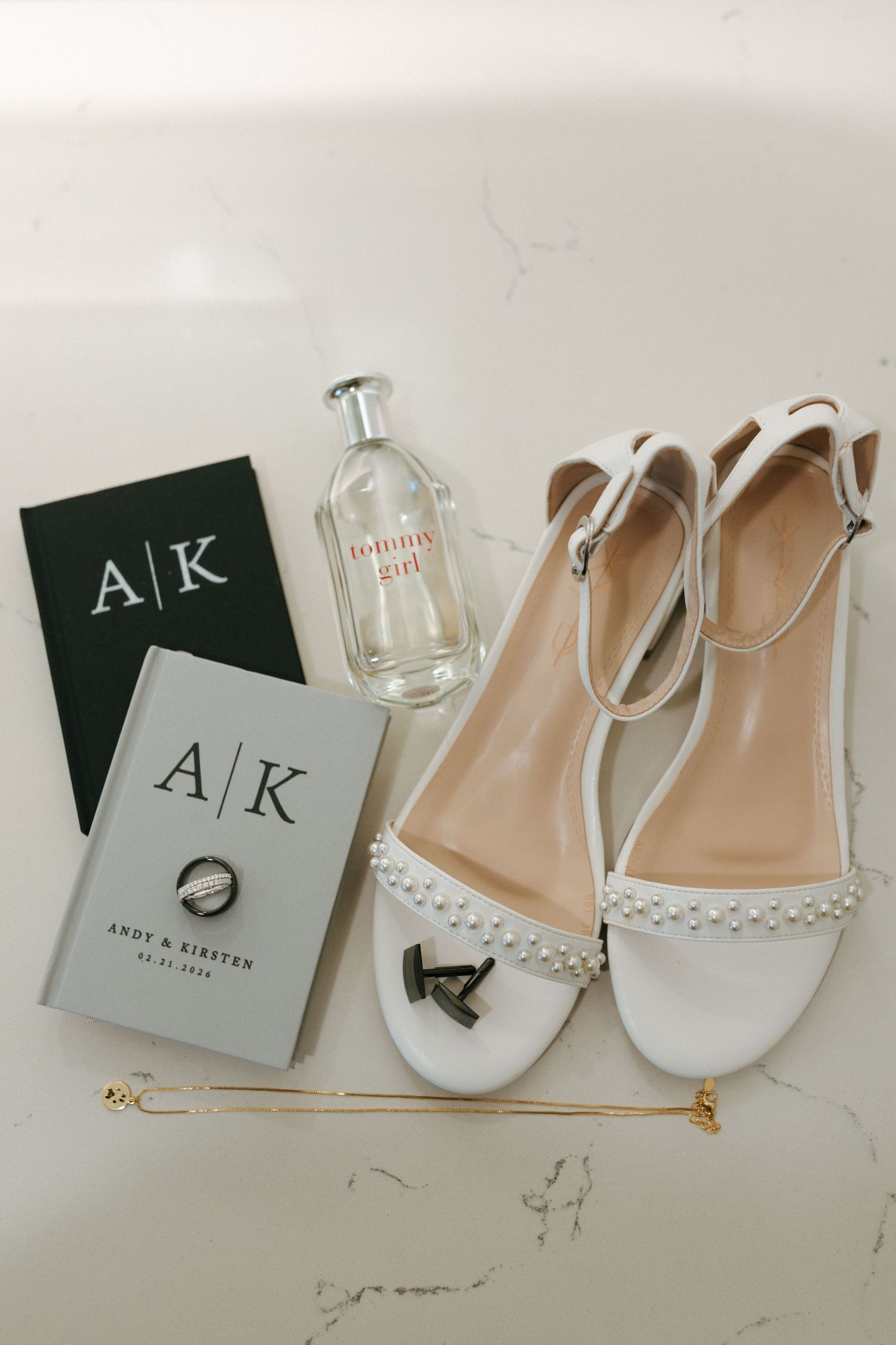 Flat lay of white pearl wedding sandals, monogrammed vow books, perfume bottle, and gold bracelet on marble.