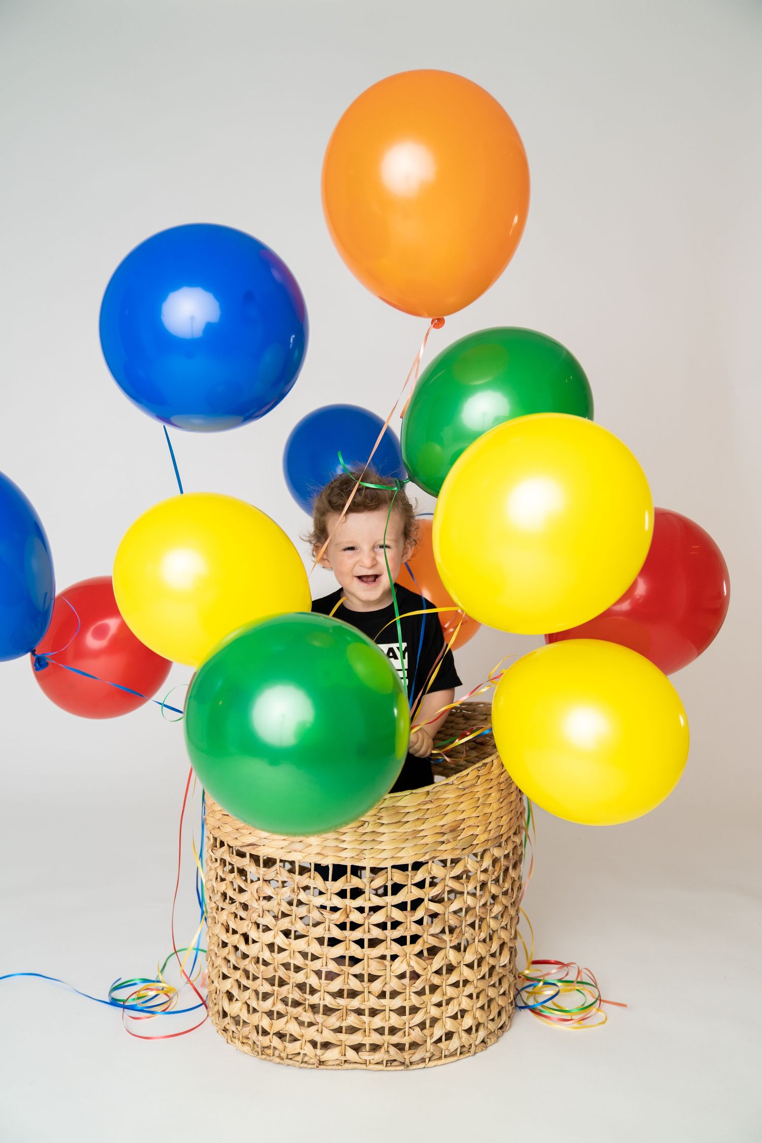 Laughing toddler boy in black shirt playing with colorful balloons inside a wicker basket.