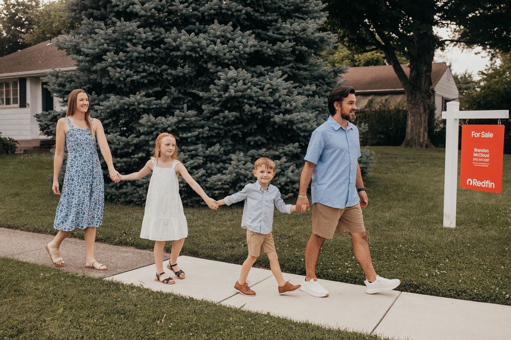 Happy family of four holding hands walking on sidewalk past a home with a for sale sign on a sunny day.