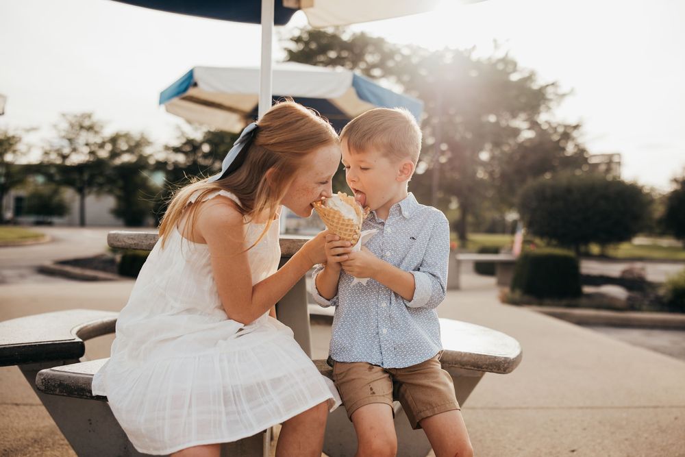 Two young children sharing an ice cream cone at an outdoor picnic table on a sunny day.