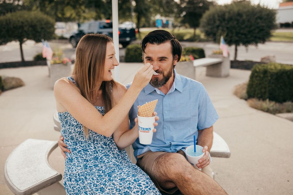 A woman feeds her bearded boyfriend french fries while sitting outdoors on a sunny day, both smiling and relaxed.