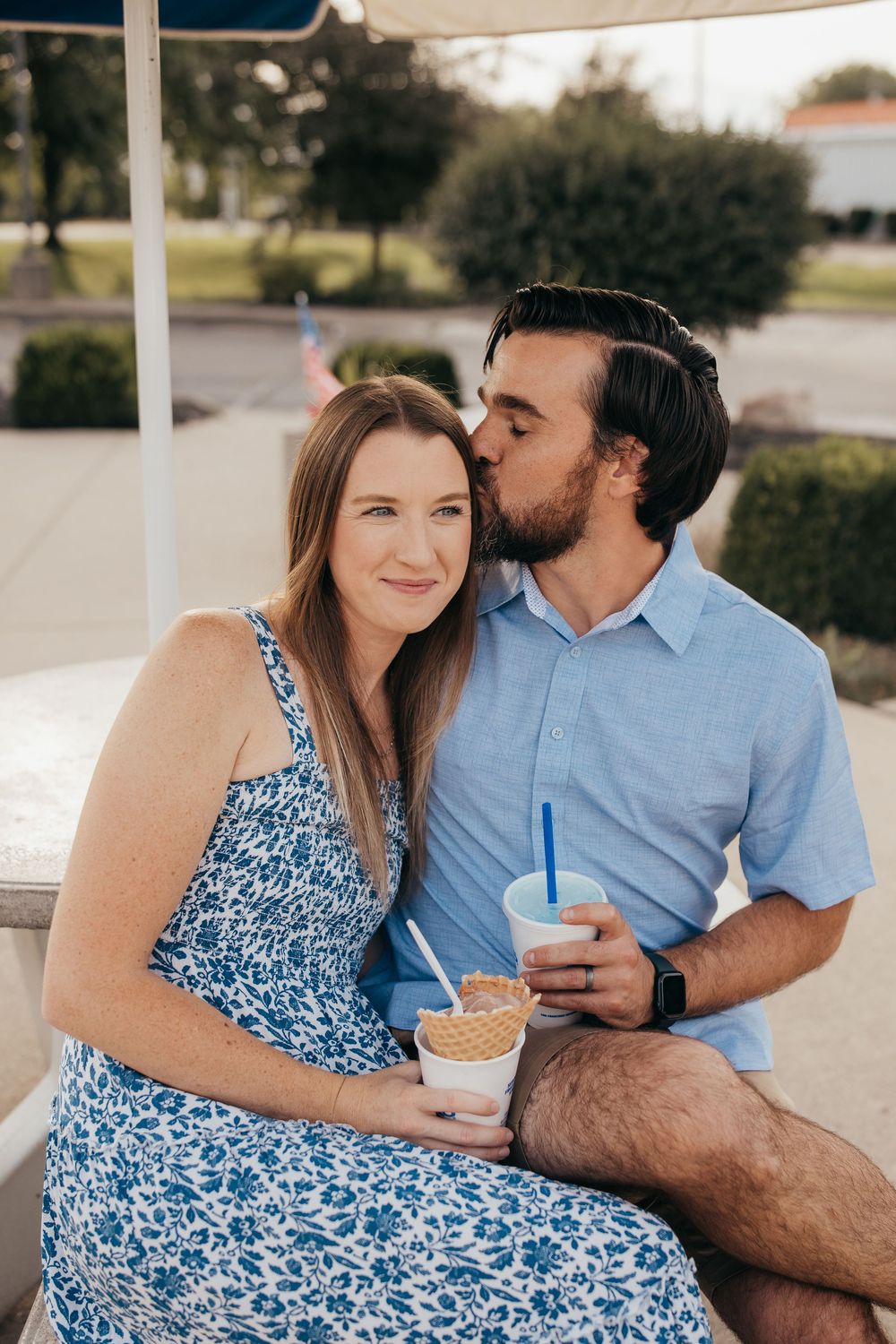 A couple shares a romantic kiss while holding frozen drinks on a bench outdoors.