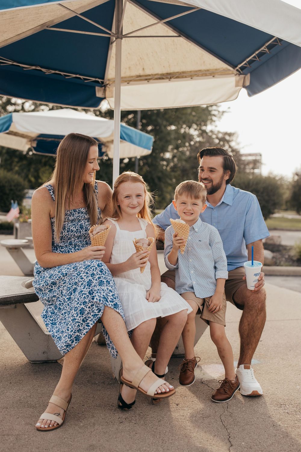 Happy family of four enjoying ice cream cones under a striped umbrella on a sunny day outdoors.