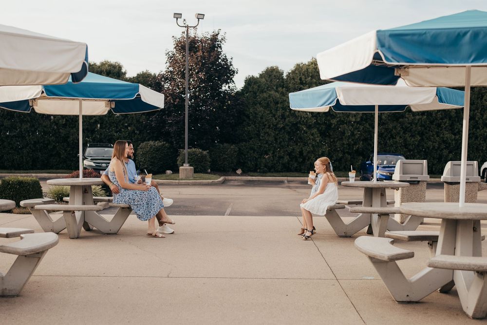 Two women socializing at outdoor picnic tables under blue and white striped umbrellas on a sunny patio.
