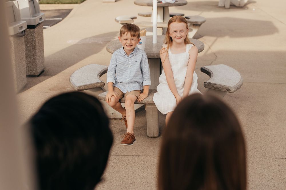 Two smiling children, a boy and girl, sit together on a stone bench at a sunny outdoor playground.