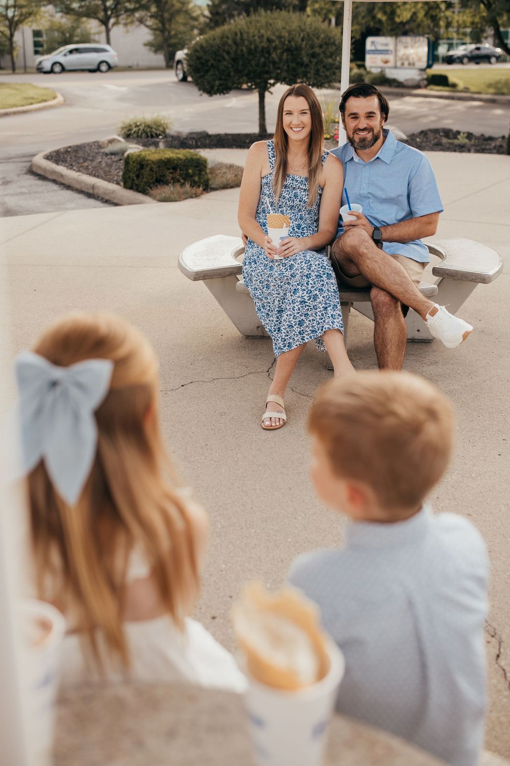 A smiling couple sits on a bench as two young children approach holding frozen treats.