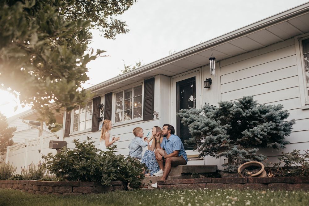 Family sitting on front porch steps of white suburban home, enjoying sunny day together in lush green yard.