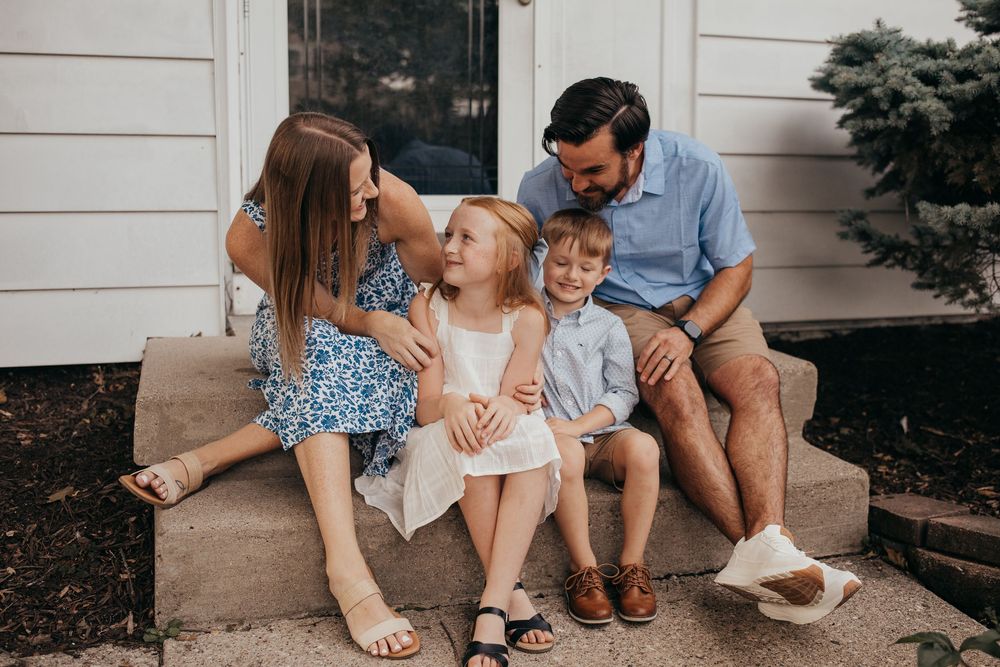 Happy family of four sitting on front porch steps, parents smiling at young children in summer outfits.