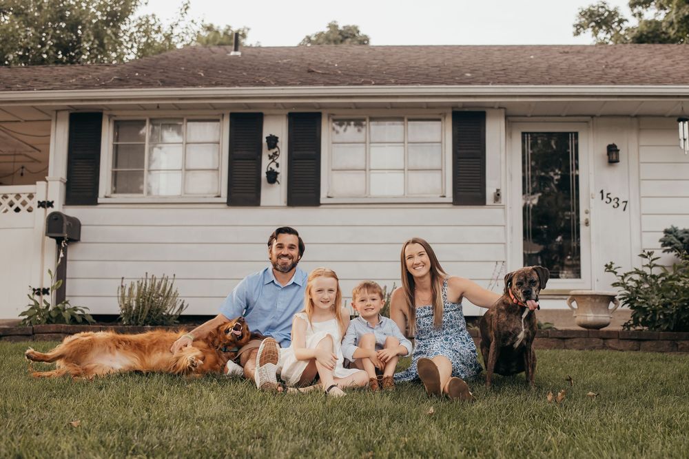 Family of four with two dogs posing on lawn in front of white ranch-style home for a portrait photo.